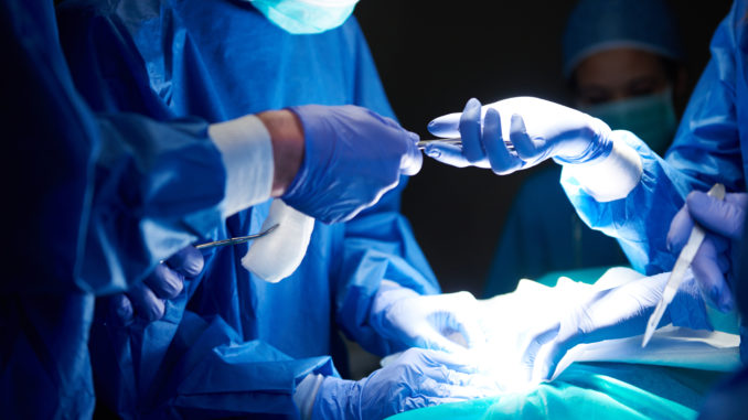 Procedimento cirúrgico em que um cirurgião passa um objeto para outro profissional. Os quatro profissionais estão com vestimentas cirúrgicas azuis e a luz emanada do local operada destaca a imagem.