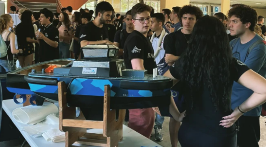 alunos apresentam a equipe na frente de calouros. Atrás deles, um barco em miniatura em cima de uma mesa